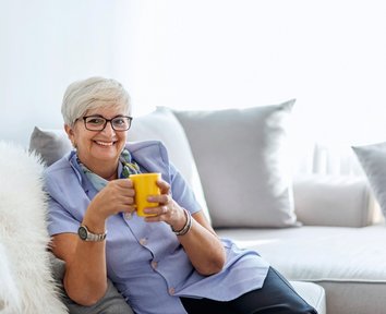 Ältere Frau mit gleber Tasse in der Hand auf dem Sofa sitzend