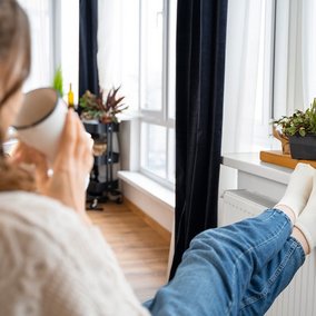 Ansicht einer sitzenden Frau von hinten, Füße auf die Heizung gelegt, Teetasse in der Hand