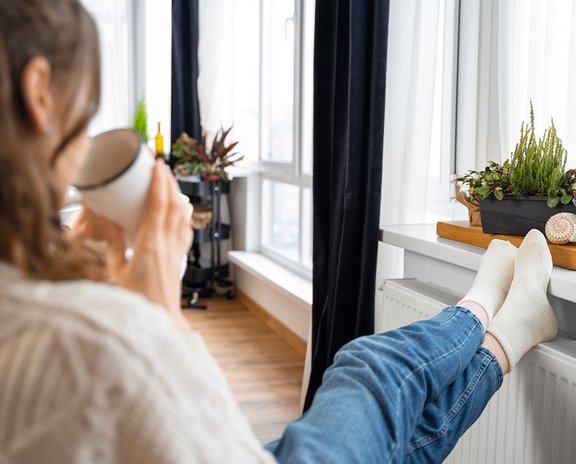 Ansicht einer sitzenden Frau von hinten, Füße auf die Heizung gelegt, Teetasse in der Hand